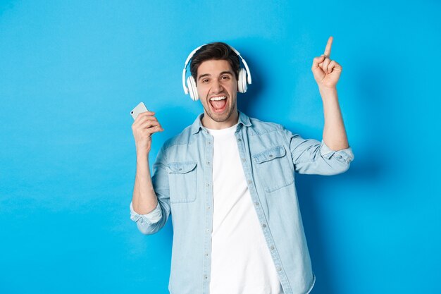 Hombre guapo alegre bailando con smartphone, escuchando música en auriculares y apuntando con el dedo hacia arriba, de pie sobre fondo azul.