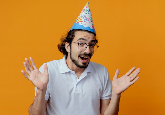 Hombre guapo alegre con gafas y gorro de cumpleaños extiende las manos aisladas sobre fondo naranja