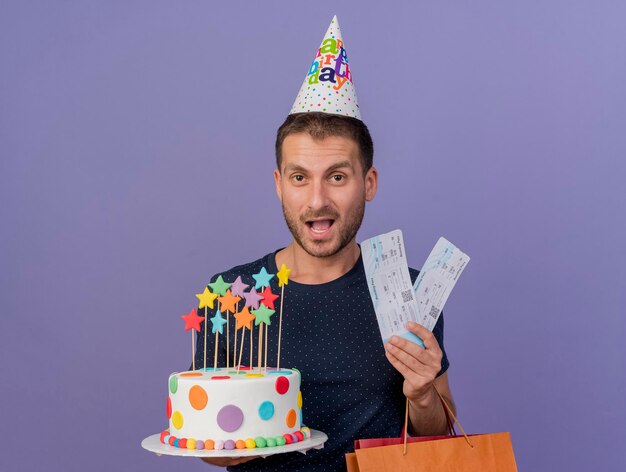 Hombre guapo alegre con gorro de cumpleaños sostiene una bolsa de compras de papel de pastel de cumpleaños y boletos de avión aislados en la pared púrpura con espacio de copia