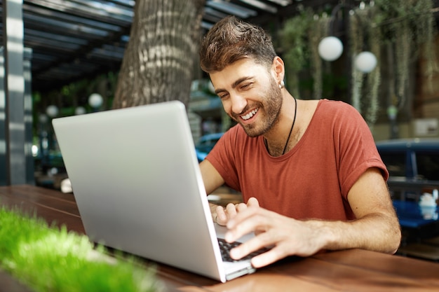 Hombre guapo alegre hablando con un amigo en línea, escribiendo en la computadora portátil y sonriendo feliz