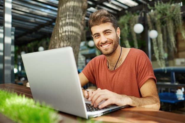 Hombre guapo alegre hablando con un amigo en línea, escribiendo en la computadora portátil y sonriendo feliz