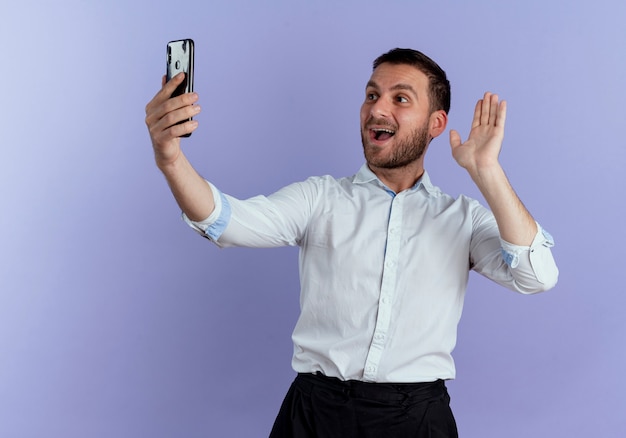 Hombre guapo alegre levanta la mano sosteniendo y mirando el teléfono aislado en la pared púrpura