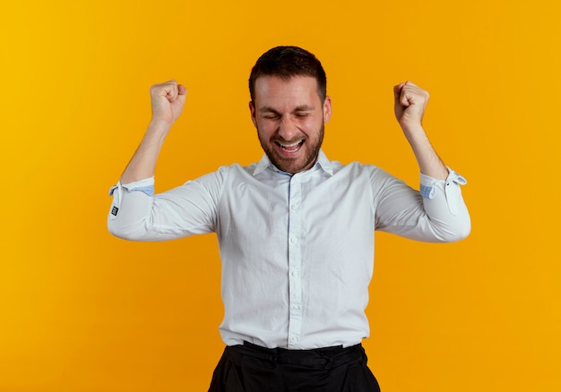 Hombre guapo alegre mantiene los puños en alto con los ojos cerrados aislados en la pared naranja