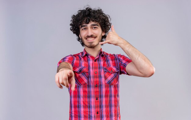 Un hombre guapo alegre con el pelo rizado en camisa a cuadros sosteniendo la mano cerca de la oreja, señal de teléfono de gesto con los dedos