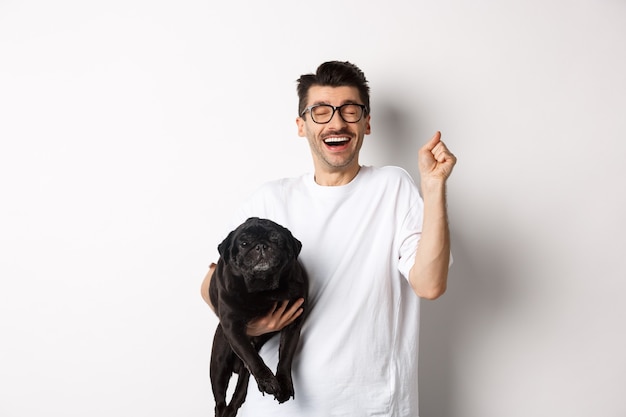 Hombre guapo alegre con perro regocijándose, celebrando la victoria. Chico feliz lleva lindo pug negro y mirando optimista, adoptando mascota, de pie sobre fondo blanco.