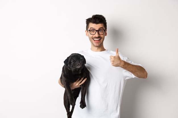 Hombre guapo alegre que sostiene el perro y que muestra el signo bien, aprueba o recomienda el producto. Chico hipster lleva lindo pug negro y mirando satisfecho, fondo blanco.