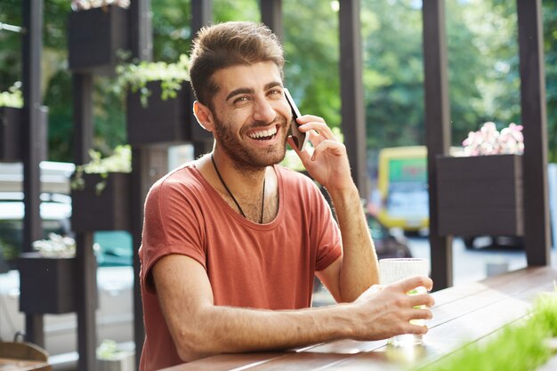 Hombre guapo alegre riendo y sonriendo mientras habla por teléfono móvil desde la cafetería al aire libre