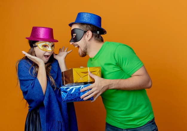 Hombre guapo alegre con sombrero azul con máscara de ojos de mascarada sosteniendo cajas de regalo mirando a una niña sorprendida con sombrero rosa y máscara de ojos de mascarada levantando las manos mirando cajas