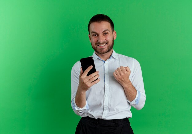 Hombre guapo alegre sostiene el teléfono mantiene el puño mirando aislado en la pared verde