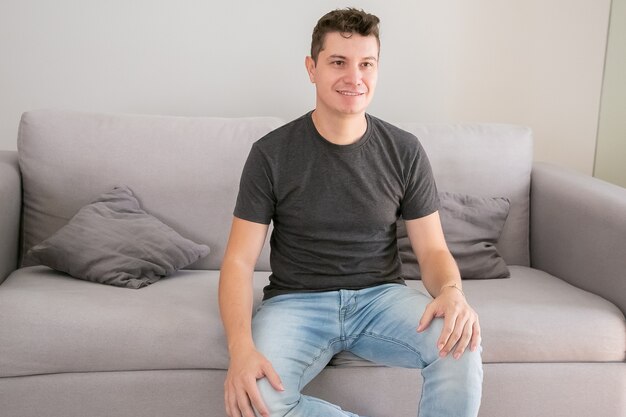 Hombre guapo alegre vestido con jeans y camiseta casual, sentado en el sofá en casa, manteniendo las manos en el regazo, mirando a otro lado y sonriendo. Concepto de retrato masculino