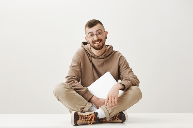 Hombre guapo amable sentarse en el suelo con el portátil y sonriendo