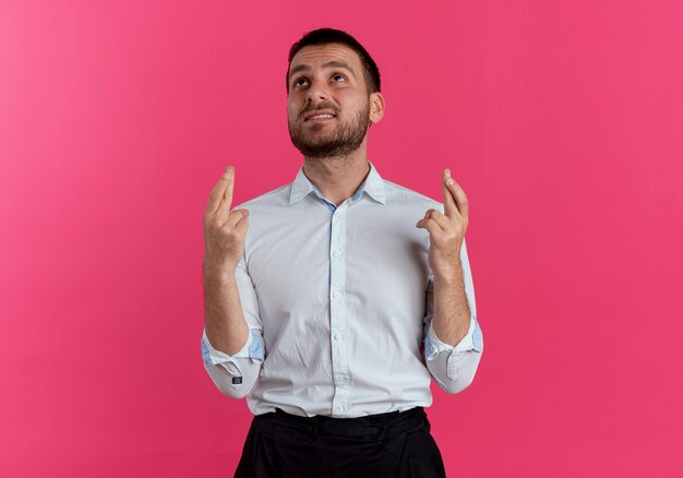 Hombre guapo ansioso cruza los dedos mirando hacia arriba aislado en la pared rosa