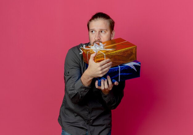 Hombre guapo ansioso sosteniendo cajas de regalo mirando al lado aislado en la pared rosa