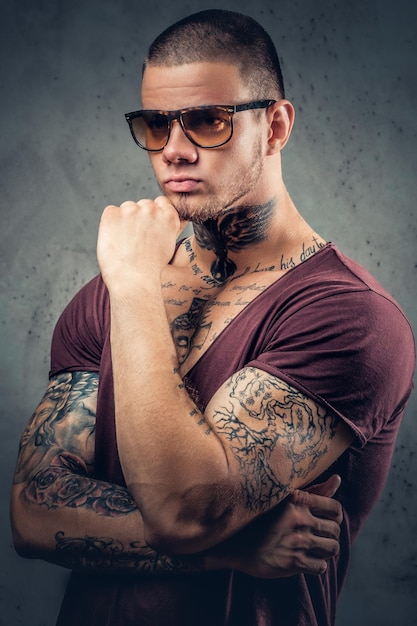 Hombre guapo y atlético con gafas de sol con tatuajes en los brazos y el cuello posando sobre un fondo artístico gris en un estudio.