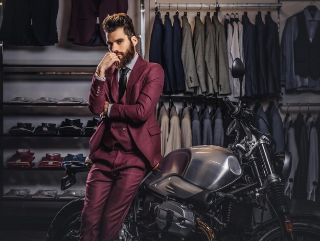 Hombre guapo con barba elegante y cabello vestido con traje rojo vintage posando cerca de una moto deportiva retro en la tienda de ropa para hombres.