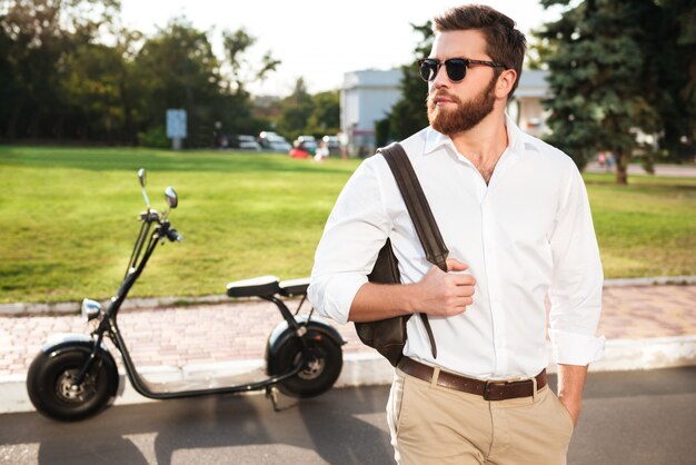 Hombre guapo con barba en gafas de sol posando al aire libre con moto moderna en el fondo