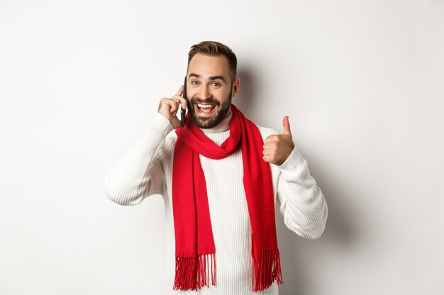 Hombre guapo con barba hablando por teléfono, mostrando los pulgares hacia arriba en señal de aprobación, como algo, confirmar el pedido, de pie sobre fondo blanco.