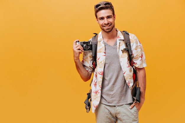 Hombre guapo con barba pelirroja en camisa blanca y camiseta a cuadros sonriendo y sosteniendo la cámara en la pared naranja aislada