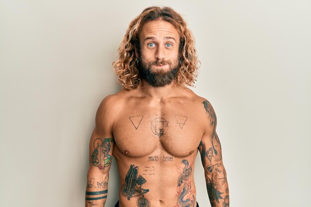 Hombre guapo con barba y pelo largo de pie sin camisa mostrando tatuajes hinchando las mejillas con cara graciosa. boca inflada de aire, expresión loca.