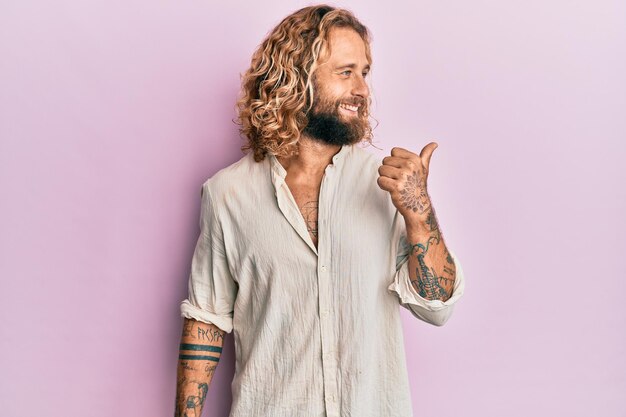 Hombre guapo con barba y pelo largo con ropa informal apuntando con el pulgar hacia el lado sonriendo feliz con la boca abierta