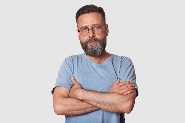 Foto gratuita hombre guapo con barba de pie con los brazos cruzados aislado en blanco, se ve serio, vistiendo camiseta gris casual y gafas, hombre posando. concepto de personas