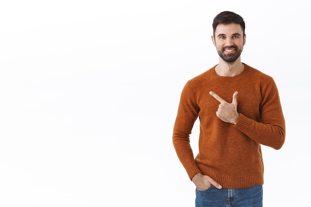 Hombre guapo con barba, propietario de la empresa que invita a apoyar su puesta en marcha, señalar con el dedo a la izquierda, mostrar información sonriendo complacido, recomendar hacer clic y averiguar