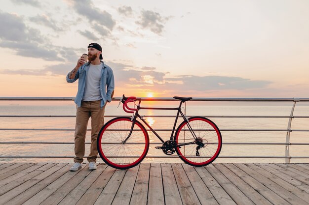 Hombre guapo con barba que viaja con bicicleta en el amanecer de la mañana junto al mar tomando café, viajero de estilo de vida activo y saludable