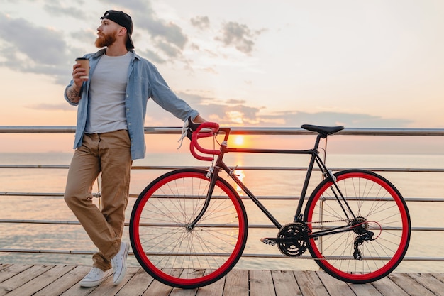 Hombre guapo con barba que viaja con bicicleta en el amanecer de la mañana junto al mar tomando café, viajero de estilo de vida activo y saludable
