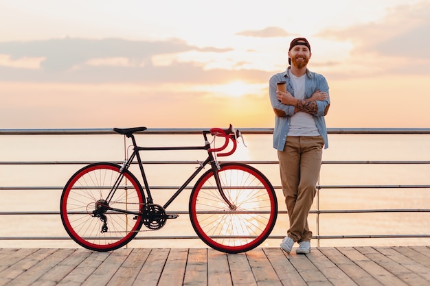 Hombre guapo con barba que viaja con bicicleta en el amanecer de la mañana junto al mar tomando café, viajero de estilo de vida activo y saludable