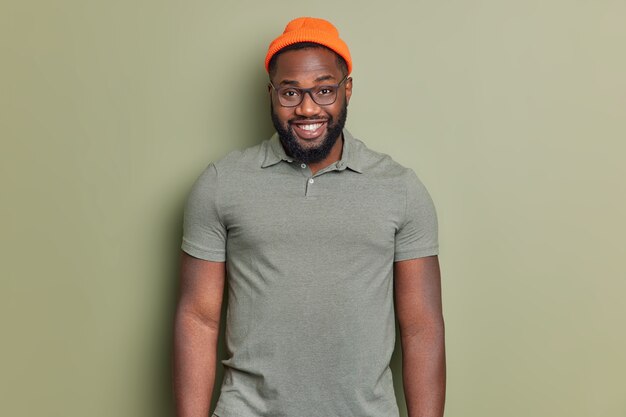 Hombre guapo con barba se siente feliz viste un sombrero naranja y una camiseta aislada sobre una pared de color caqui