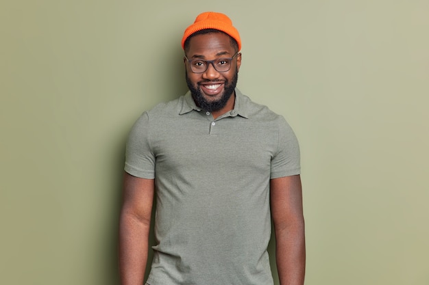 Hombre guapo con barba se siente feliz viste un sombrero naranja y una camiseta aislada sobre una pared de color caqui