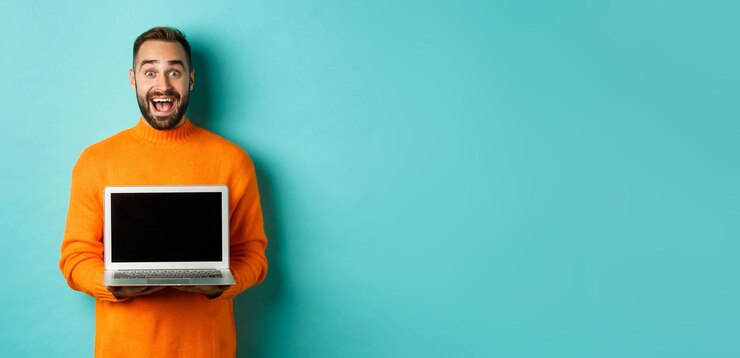 Foto gratuito hombre guapo con barba en suéter naranja que muestra la pantalla del portátil que muestra la promoción de pie sobre la luz