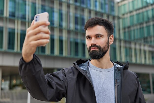 Hombre guapo con barba toma selfie a través de teléfono inteligente viste chaqueta negra paseos en poses del centro frente a la construcción de la ciudad moderna disfruta de tiempo libre. Concepto de tecnología y estilo de vida urbano de personas.