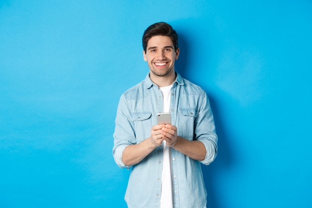 Hombre guapo con barba en traje casual sonriendo a la cámara, comprobando el teléfono inteligente, de pie contra el fondo azul