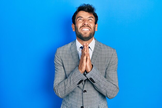 Hombre guapo con barba con traje de negocios y corbata rogando y rezando con las manos juntas con expresión de esperanza en la cara muy emotivo y preocupado rogando