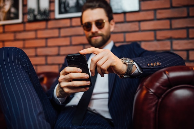 Hombre guapo con barba vestido con traje sentado en el sofá de cuero con smartphone. Confort y relajación.