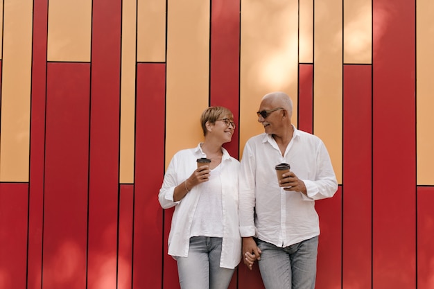 Hombre guapo con camisa blanca cogidos de la mano con dama rubia en blusa ligera y anteojos con taza de té en rojo y naranja.