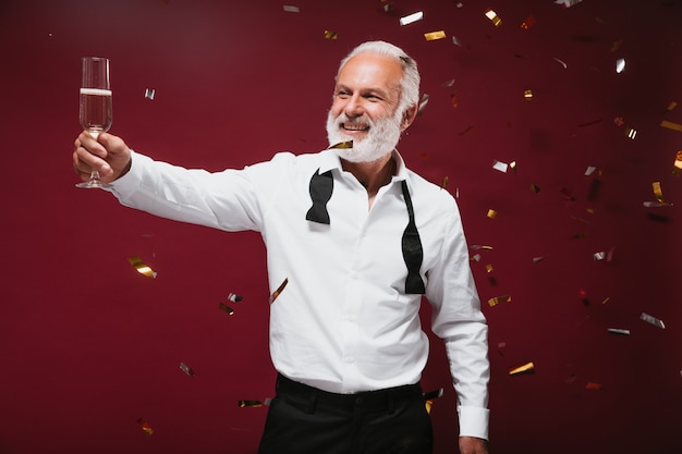 Foto gratuita hombre guapo con camisa blanca sostiene copa de champán y posa en la pared de color burdeos