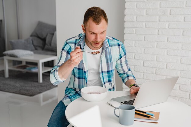 Hombre guapo en camisa desayunando en casa en la mesa trabajando en línea en la computadora portátil desde casa