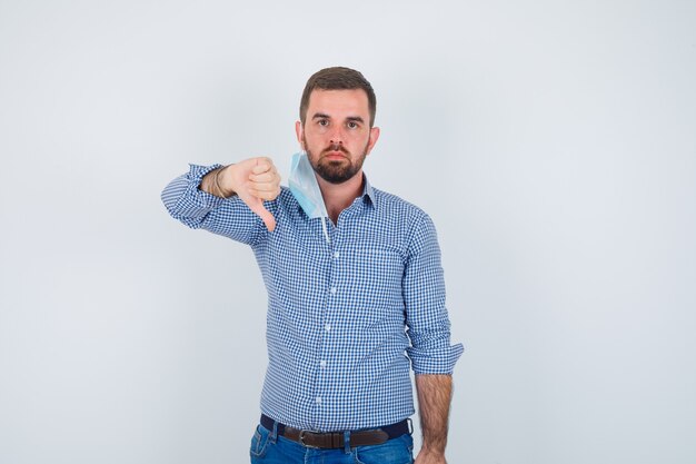 Hombre guapo en camisa, jeans, máscara quitándose la máscara, mostrando el pulgar hacia abajo y mirando disgustado, vista frontal.