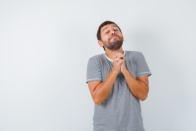 Hombre guapo en camiseta manteniendo los dedos entrelazados y mirando esperanzado, vista frontal.