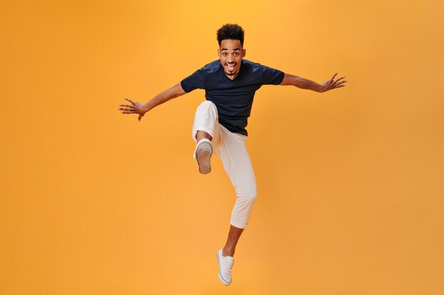 Hombre guapo con camiseta negra sonríe y salta sobre fondo naranja Chico genial con pantalones blancos moviéndose sobre un fondo aislado