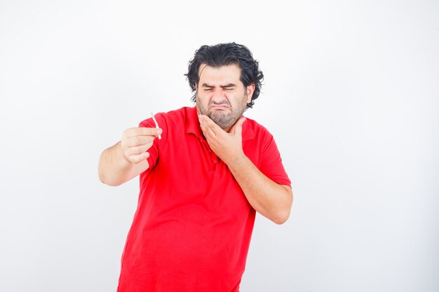 Hombre guapo en camiseta roja sosteniendo un cigarrillo, sosteniendo la mano en el cuello, haciendo muecas y mirando disgustado, vista frontal.