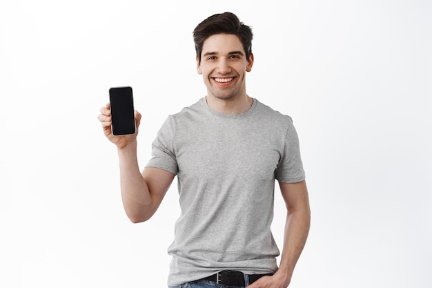 Hombre guapo caucásico muestra la pantalla del teléfono móvil, sonriendo y dando recomendación de la aplicación, de pie en camiseta contra el fondo blanco.
