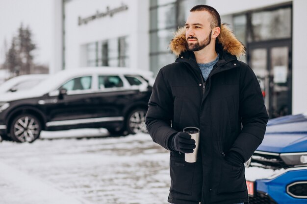 Hombre guapo en chaqueta de abrigo de pie en coche cubierto de nieve y tomando café