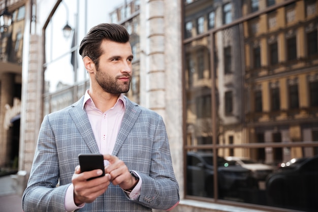 Hombre guapo en una chaqueta de pie y sosteniendo teléfono móvil