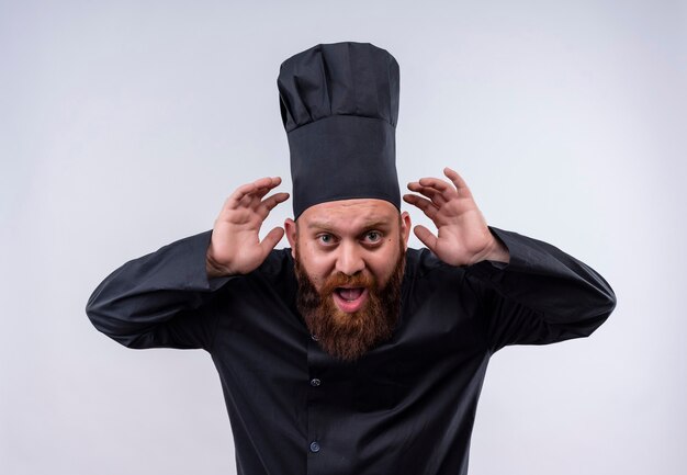 Un hombre guapo chef barbudo en uniforme negro con las manos en los oídos tratando de escuchar algo en una pared blanca