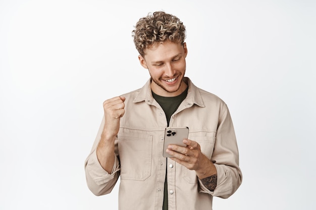 Hombre guapo complacido animando mientras mira su teléfono móvil Lograr el objetivo en la aplicación en el teléfono inteligente y celebrando de pie sobre fondo blanco