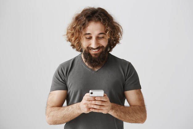 Hombre guapo complacido enviando mensajes de texto en el teléfono inteligente, mirando el teléfono móvil encantado