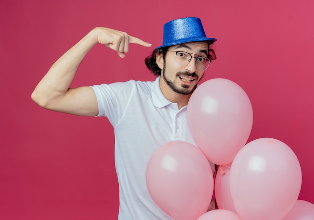 Hombre guapo complacido con gafas y sombrero azul sosteniendo globos y puntos al lado aislado sobre fondo rosa